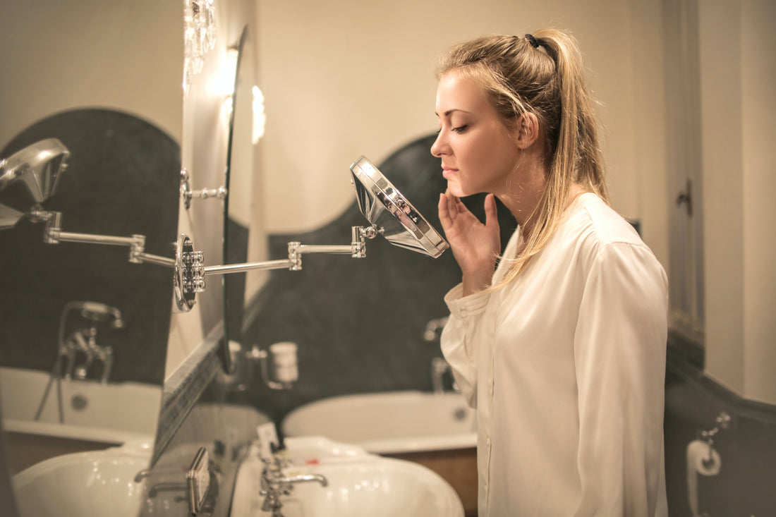 Blonde woman wearing white pajamas looking at skin in a wall-mounted mirror