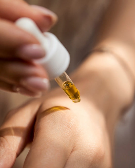 A feminine hand applies a golden-toned skin nutrition oil to her other hand from a white banded glass dropper.