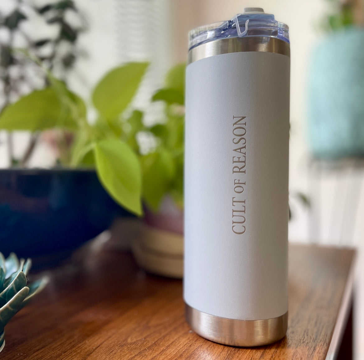 Light Grey Insulated 12oz Travel Tumbler with laser-etched Cult of Reason Logo on table with plant