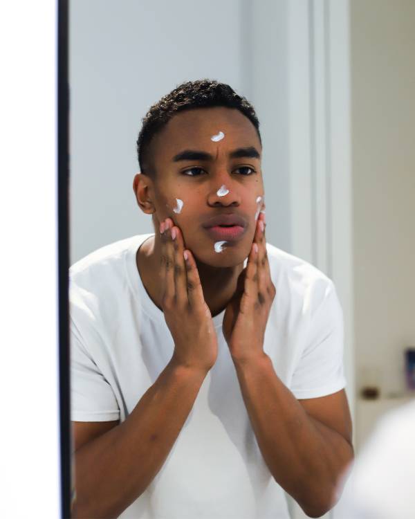 A black man in a white t-shirt has applied five dots of white cream to the five areas of his face. His hands rest on his lower cheeks while he looks into a mirror.