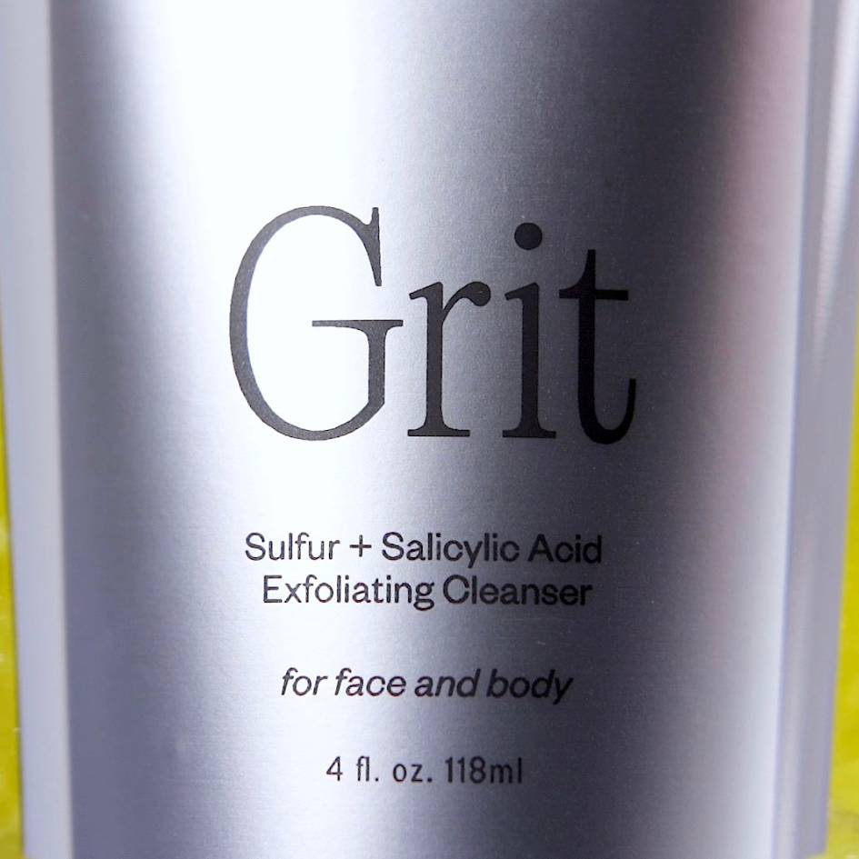 Cult of Reason Grit has a silver label with serif logo. A dramatic shadow falls across the label and slivers of chartreuse color can be seen behind the skincare tube.