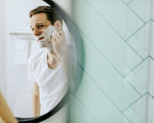 A Caucasian man wearing a white v-neck tshirt shaves in a mirror. 