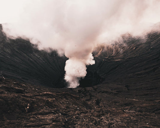 A black volcano spews white smoke into the air during an eruption.